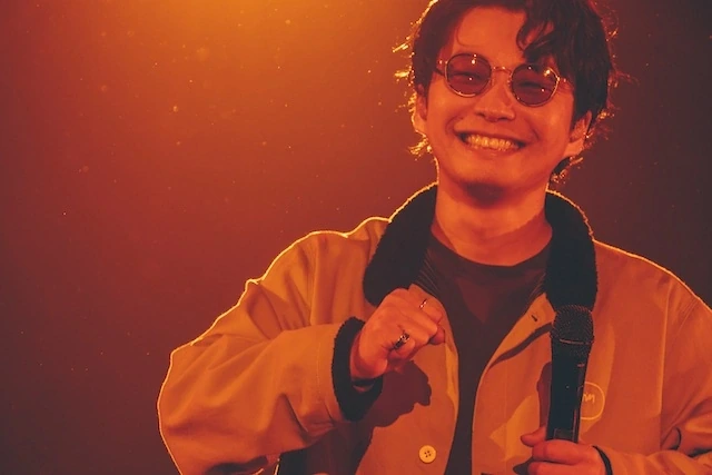 星野源さんがライブ「Reassembly」で黄色のジャケットにBJ CLASSICのPREM-137CWラウンドサングラスを着用して笑顔を見せる姿